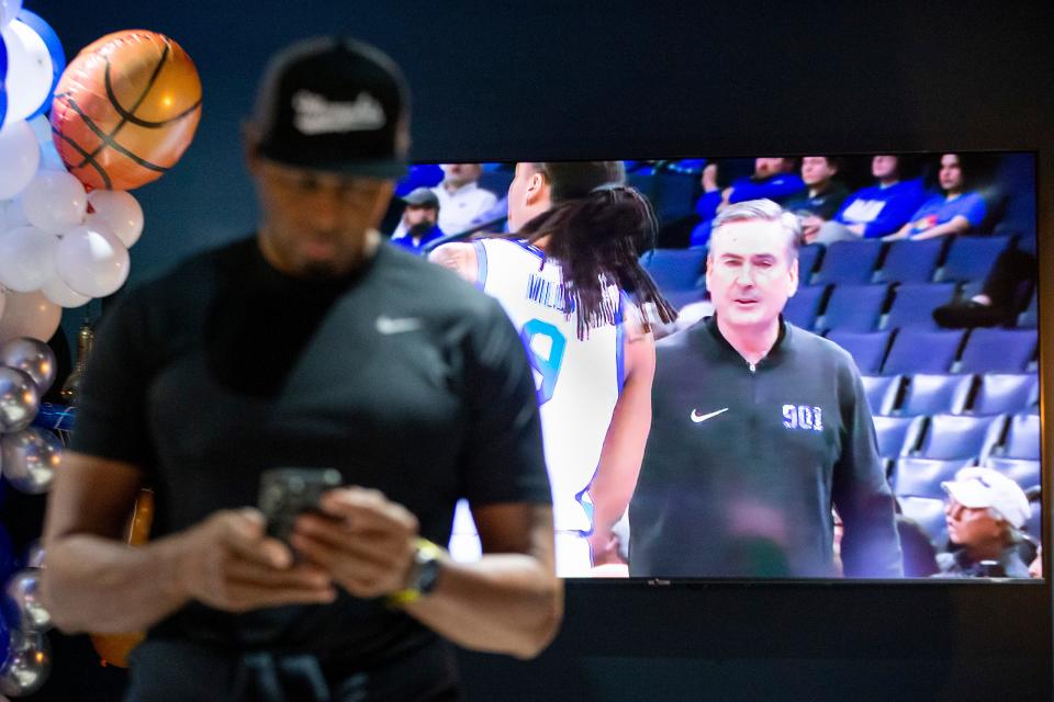 Memphis basketball head coach Penny Hardaway checks his phone as assistant coach Rick Stansbury, who is acting as head coach through Hardaway’s three-game NCAA suspension, is seen on the television behind him as Hardaway watches Memphis play Alabama State from a private room at Owners Box Sports Grill in Lakeland, Tenn., on Friday, November 17, 2023.