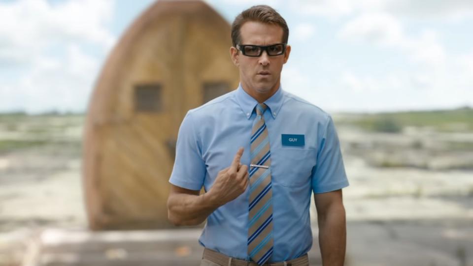 Ryan Reynolds in a blue shirt and glasses points at himself