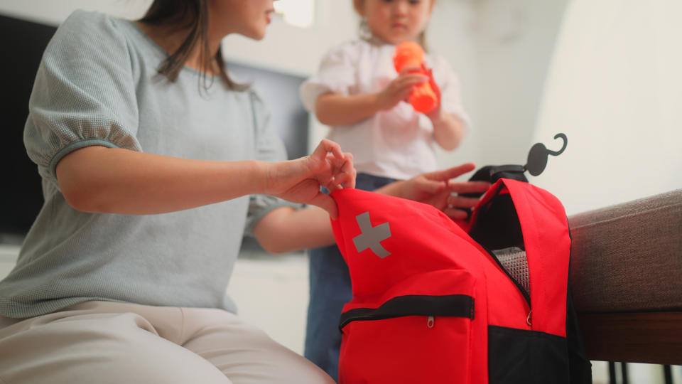 A person packs a red emergency kit while a child holds a toy. The scene suggests preparation for emergencies at home