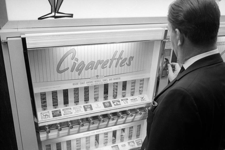 A man stands in front of a cigarette vending machine, preparing to make a selection. The machine displays various cigarette brands