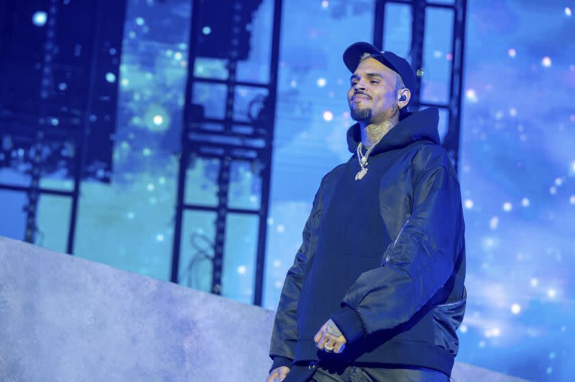 Man, Chris Brown, dressed on blue lit stage in black hat, jacket, hoodie and pants, smiling to audience