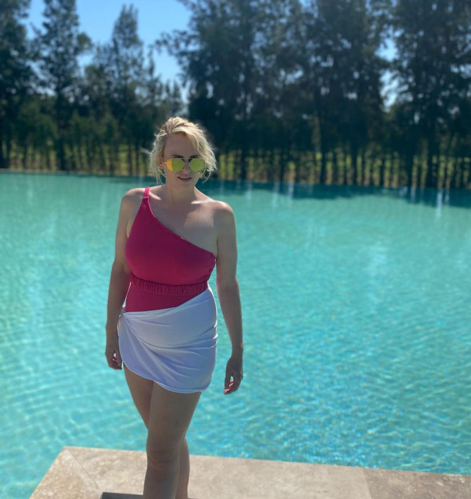 Rebel Wilson poses in front of a pool, wearing a one shoulder pink swimsuit with a white sarong tied around her waist.