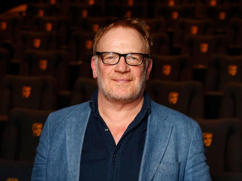Mark Lewis Jones smiling in an auditorium with Bafta-branded charis while wearing a pair of glasses and a blue blazer.