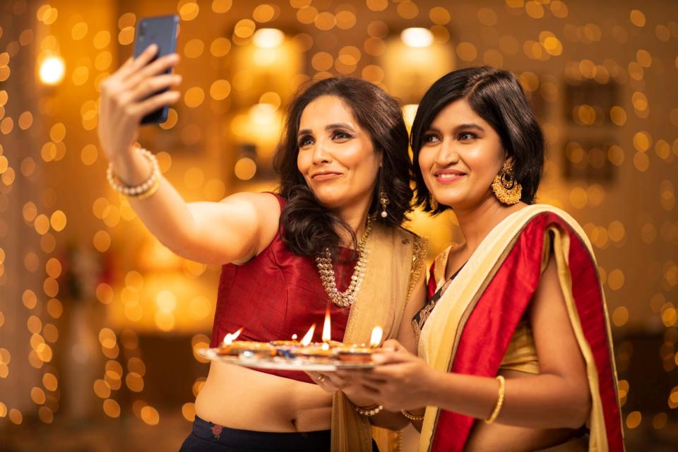 two-women-in-saris-take-a-selfie-in-front-of-string-curtain-lights-at-diwali-party