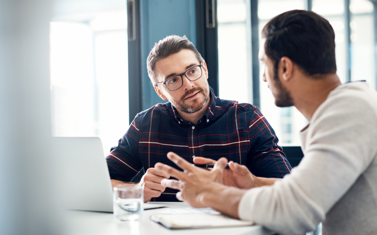 Focus on two men negotiating at work, sitting at a table with a laptop, sunlight coming through windows