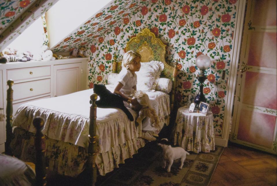 Cosima Ungaro and her Jack Russell Terrier dog, Olli, sit in her floral bedroom in the home she shares with her father, fashion designer Emanuel Ungaro and her mother, Laura Ungaro (nee Bernabei). The floral wallpaper is a rose chintz originally made for Emanuel Ungaro's couture collection by Rainbow, a collaborator. The bed linens and side table skirt are of a ruffled white floral; the white cabinets are built in under the dormer window; a lamp with a large glass globe sits on the side table. Cosima sits with two stuffed animal toys.