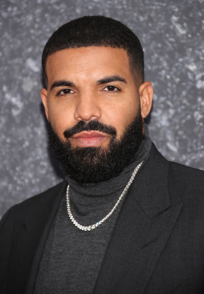 Drake wearing a black turtleneck and blazer, with a beaded necklace, posing against a textured backdrop