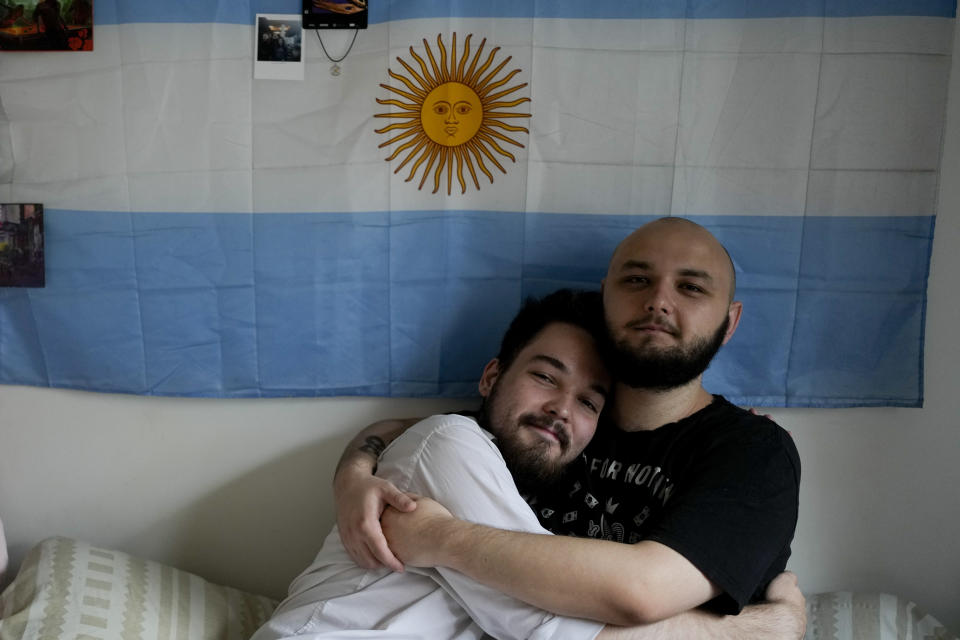 Los rusos Dmitri Yarin, a la izquierda, y su pareja Nikolai Shushpan, se abrazan frente a una bandera argentina en su casa en Buenos Aires, Argentina, el lunes 13 de febrero de 2023. Shuspan disfruta vivir abiertamente como una persona gay por primera vez. Shuspan dijo que en Rusia siempre había "tensión" y el temor de que algo pudiera suceder. (AP Foto/Natacha Pisarenko)