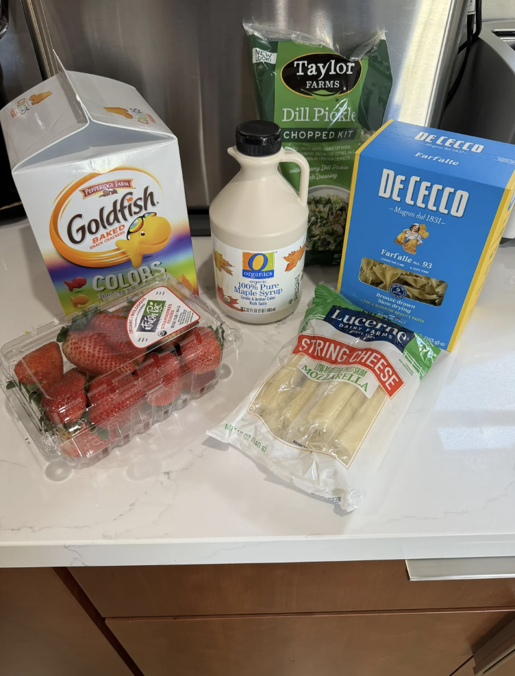 Grocery items on a kitchen counter include Goldfish crackers, Taylor Farms dill pickle chopped kit, maple syrup, De Cecco pasta, strawberries, and string cheese