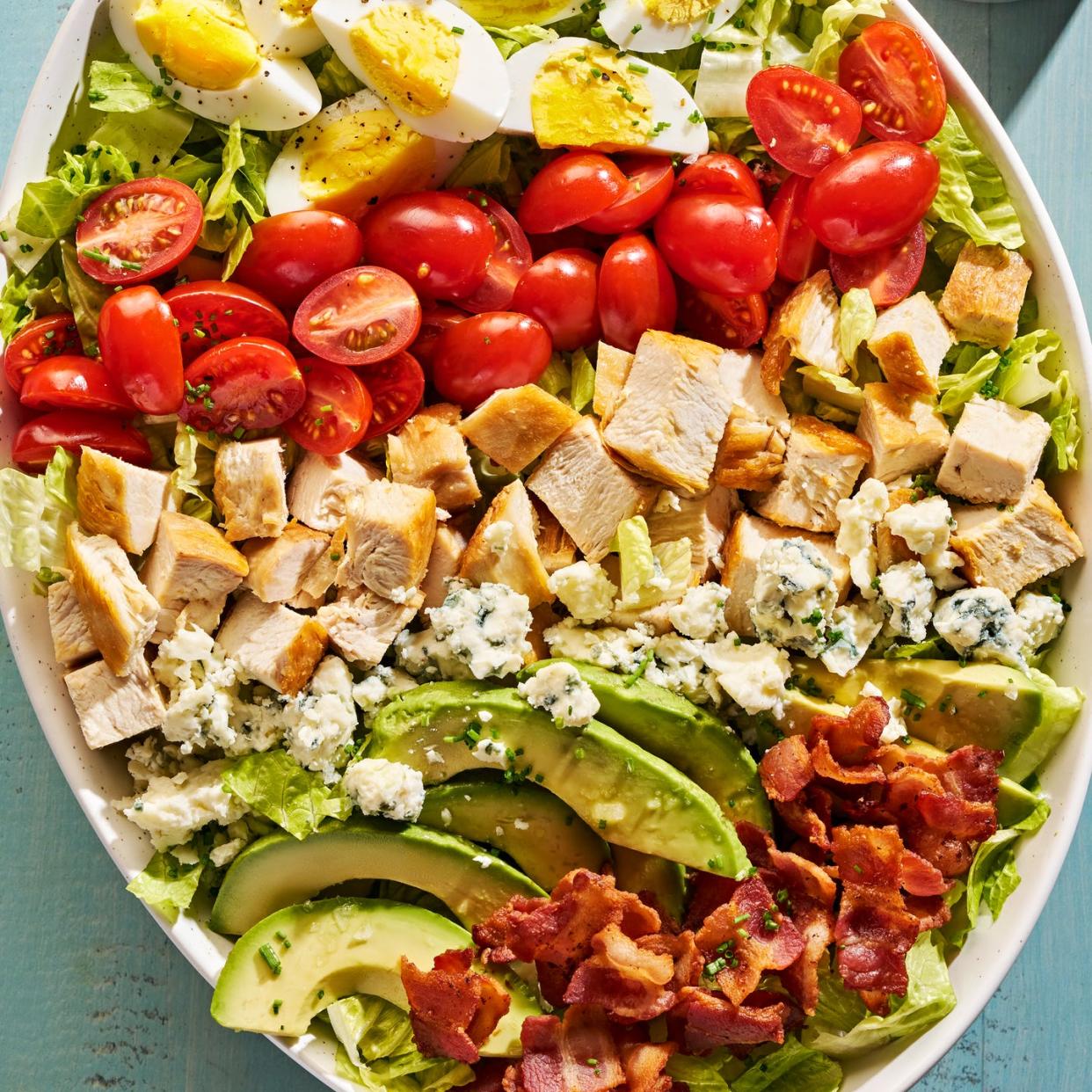 lettuce, bacon, avocado slices, hard boiled eggs, halves tomatoes, blue cheese and chicken pieces in a bowl