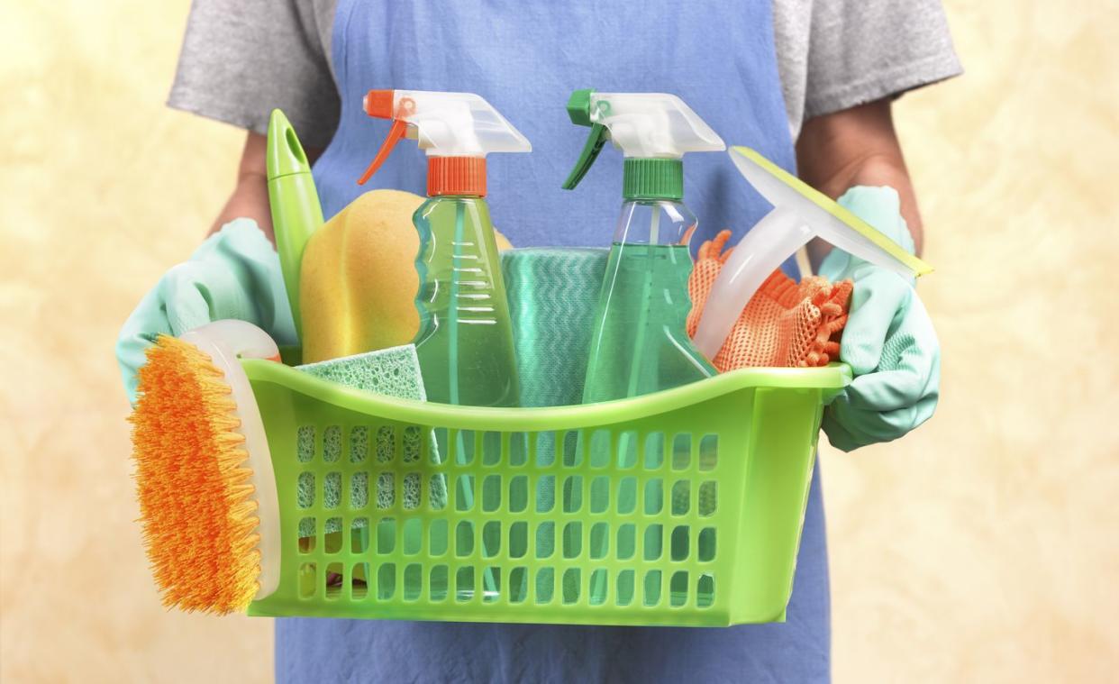 close up of a woman holding a basket containing colorful cleaning materials like squeeze rigger bottles, a brush and a sponge
