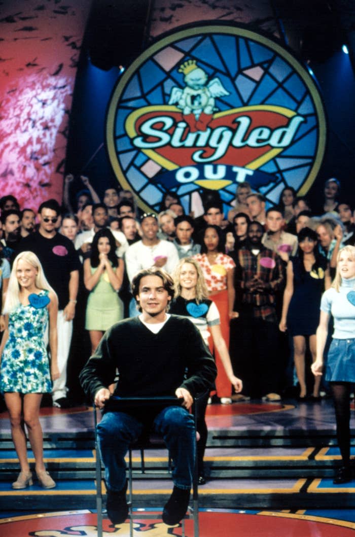 A man sitting center stage on MTV's 'Singled Out' game show with host and contestants in the background