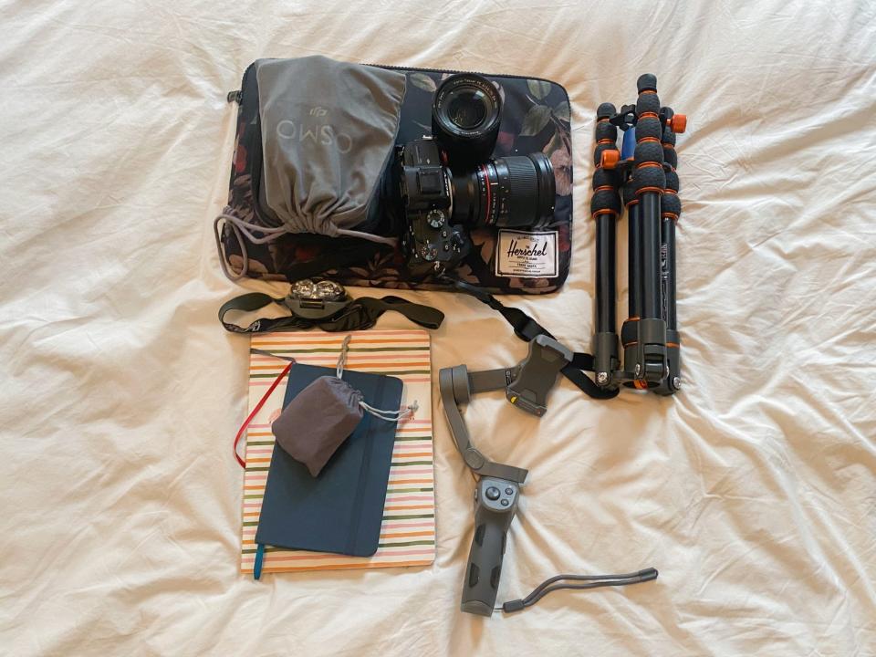 The author's work gear on a white sheet, including a black camera and lens as well as a grey pouch on top of a laptop in a black floral sleeve. To the left, there's a black tripod with orange accents folded up. Beneath all of this is two stacked notebooks and mobile photography gear.