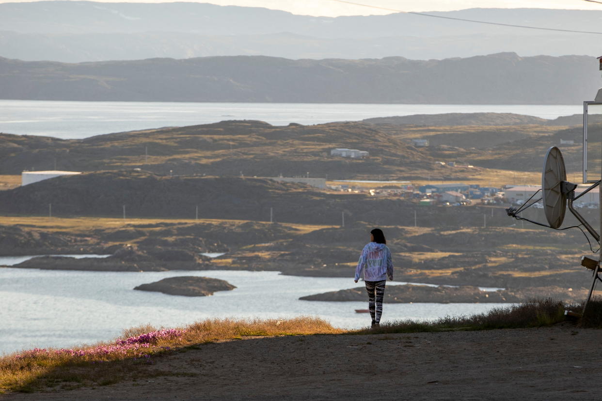 A person walks along a path on a hill overlooking a landscape of water, some buildings and distant mountains.