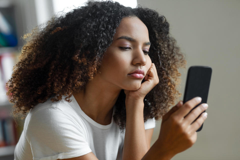 Woman looking thoughtfully at smartphone, resting chin on hand, with curly hair and white top