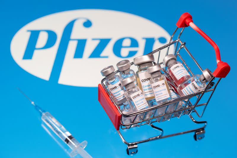 A small shopping basket filled with vials labeled "COVID-19 - Coronavirus Vaccine" and a medical sryinge are placed on a Pfizer logo