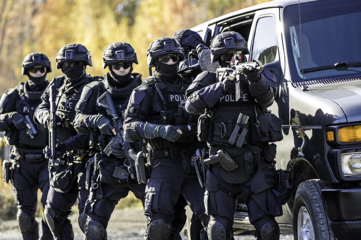 A row of six police on a SWAT team line up in battle dress, two readying their rifles and one a handgun, beside a large black van. 