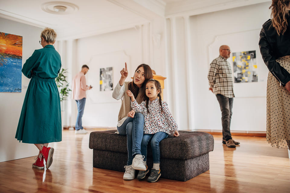 Mom and child sitting in an art gallery while the mom is pointing at something, with other visitors around