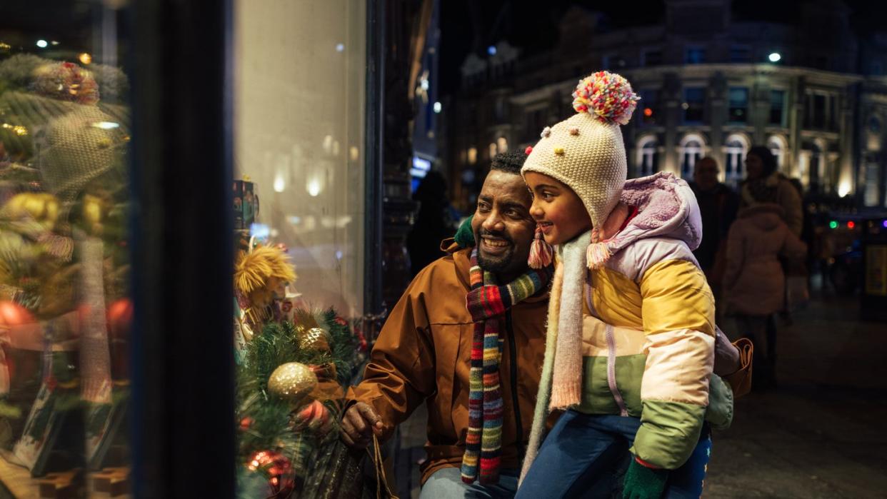 side view of a father standing outside of a store window with his young daughter, they are shopping for christmas presents and theyre wrapped up in warm clothing on a cold december night