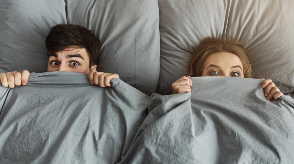 Surprised couple hiding under blanket in bed at home, covering half face, looking at camera, top view