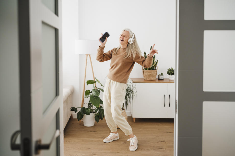 An elderly woman, wearing a brown sweater and beige pants, enjoys dancing with headphones on and holding a phone in a modern, plant-decorated room