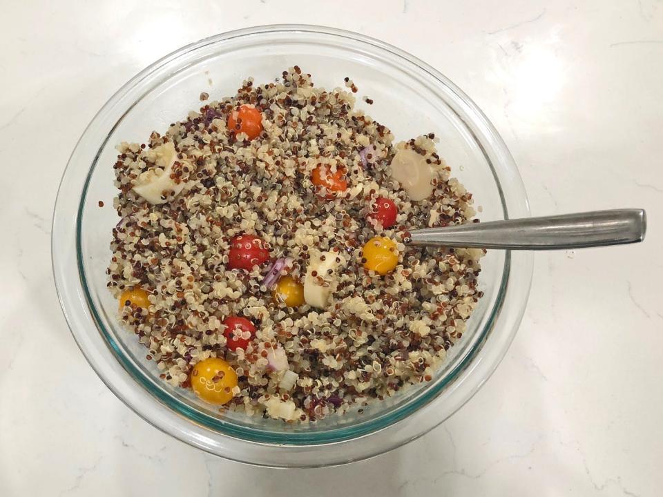 A clear bowl filled with quinoa salad, made with sliced red onions, hearts of palm, cherry tomatoes, and cooked quinoa.