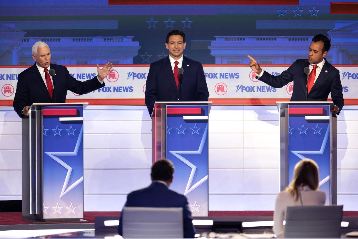 Three men grinning and gesticulating at lecterns in front of debate moderators.