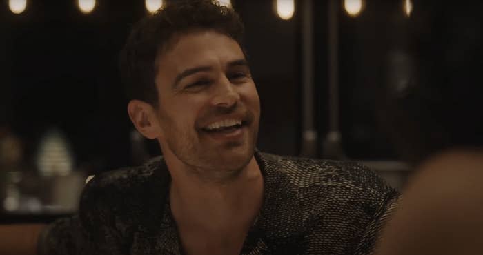 Theo James smiling at a dimly lit dinner setting. He is wearing a casual patterned shirt