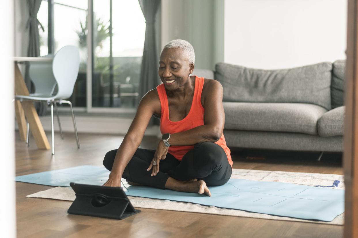 A beautiful senior woman takes an online yoga class. She is interacting with the teacher on her tablet and about to commence her class. She is wearing casual active wear and is taking the class in her lounge room. She is sitting on a blue yoga mat.