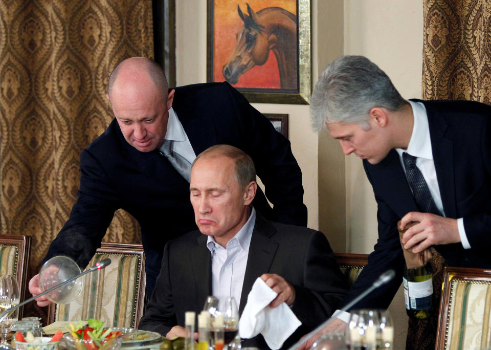 Yevgeny Prigozhin leans over Russian Prime Minister Vladimir Putin, who is seated at a table set for dinner, with a napkin in his hand, wearing a dismissive expression.