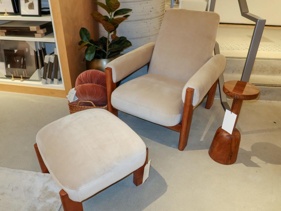 A chair with a wooden frame and beige cushions, with a similar stool in front of it. A small wooden table sits next to it.