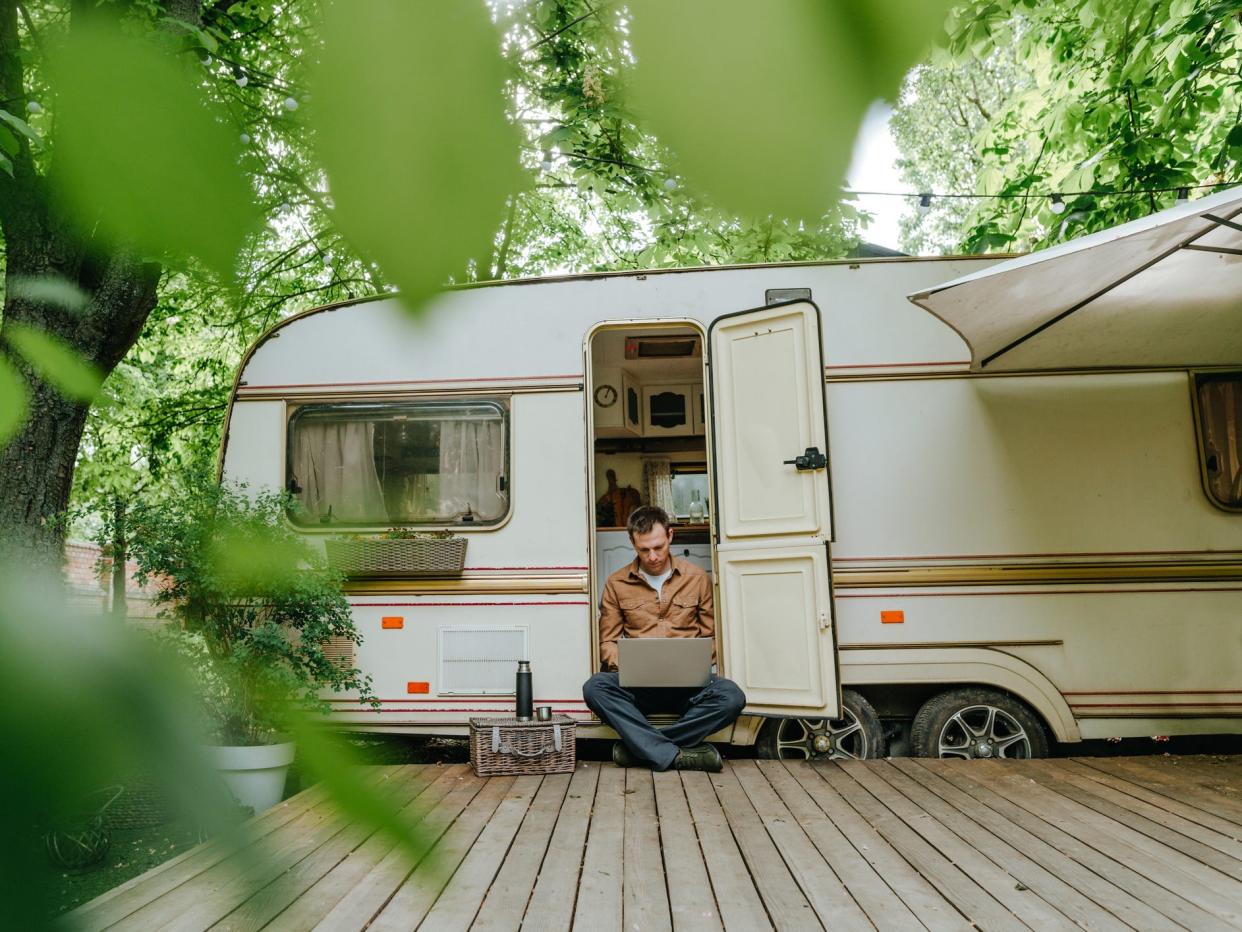 Young man drinking tea from a thermos and working on a laptop while sitting on the threshold of a camper in the forest.