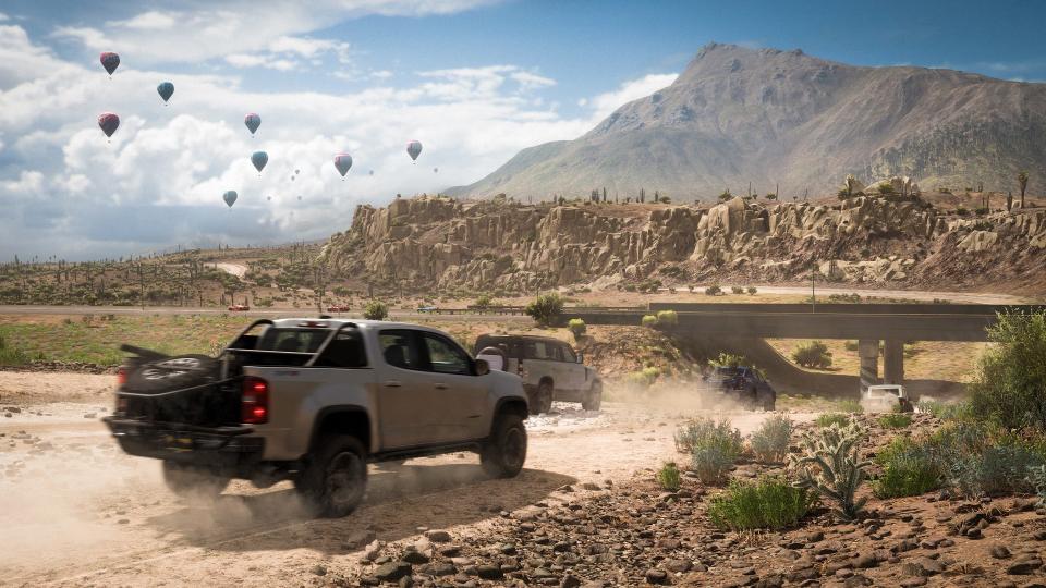 Several cars driving down a dusty desert road, craggy mountains in the distance