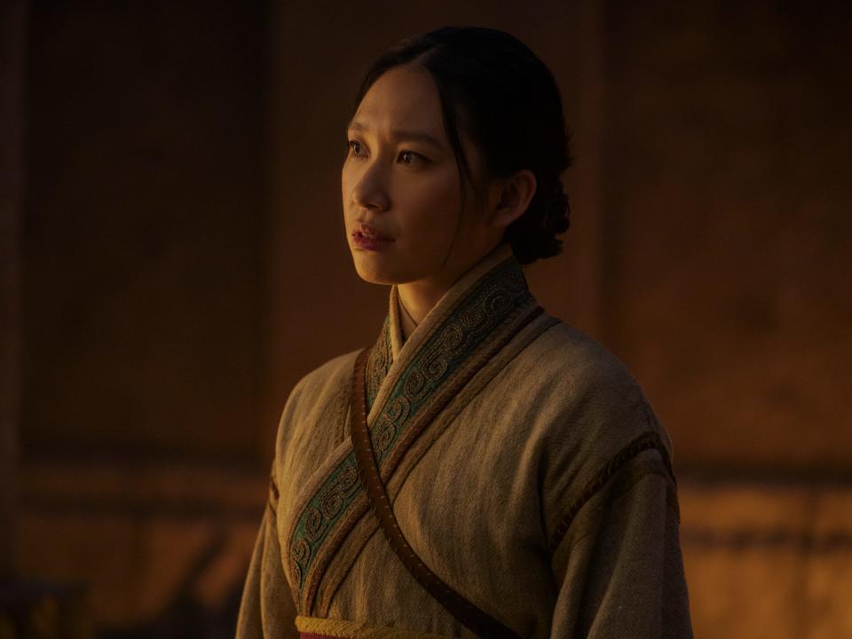 jess hong as jin cheng, wearing somewhat plain historical robes, her hair pulled back into a low bun. she's half lit from the side from a warm light source