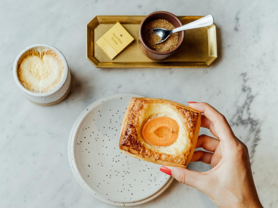 A hand holding a baked good over a plate next to coffee and sugar.