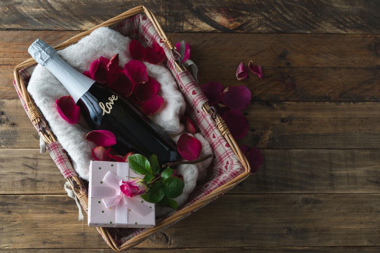 Valentine's Day. Gift basket with champagne bottle with LOVE inscription on the bottle and gift box with rose. Decoration with rose and red petals on wooden background. Copy space. Top view.