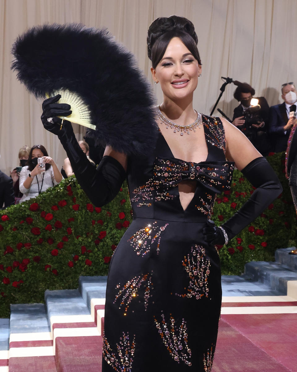 Woman in a cutout gown with bead detailing, holding a feathered fan, at a gala event