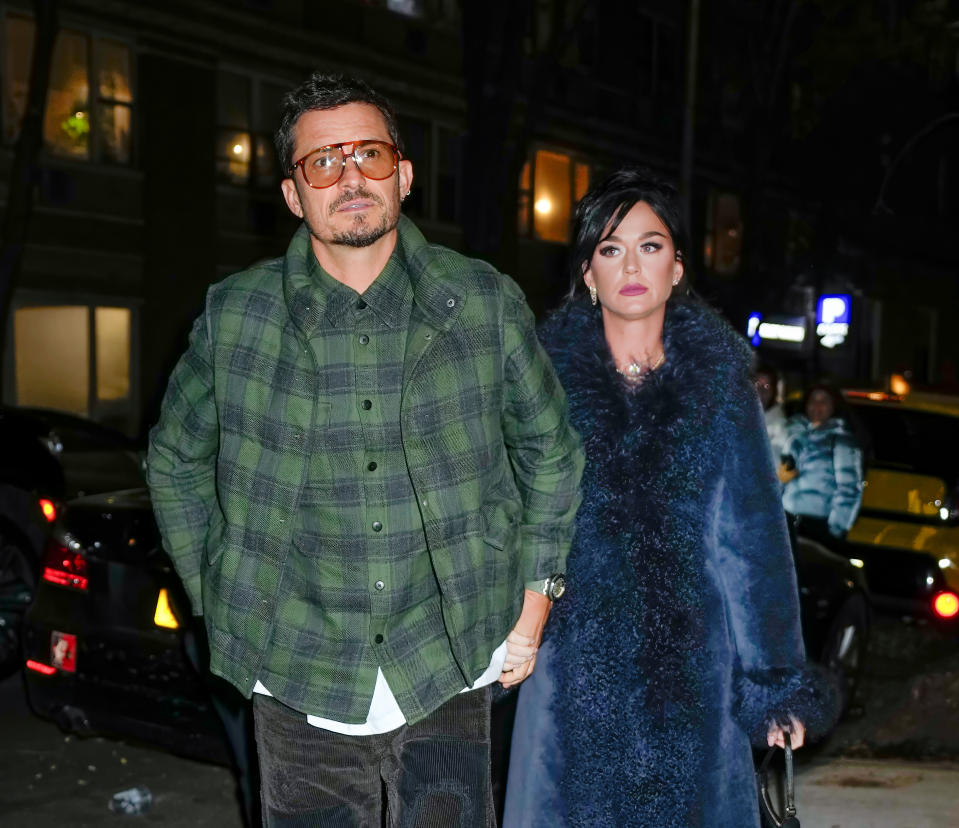 Orlando Bloom and Katy Perry hold hands while walking outside. Orlando wears a green checkered jacket and sunglasses. Katy is in a fur coat