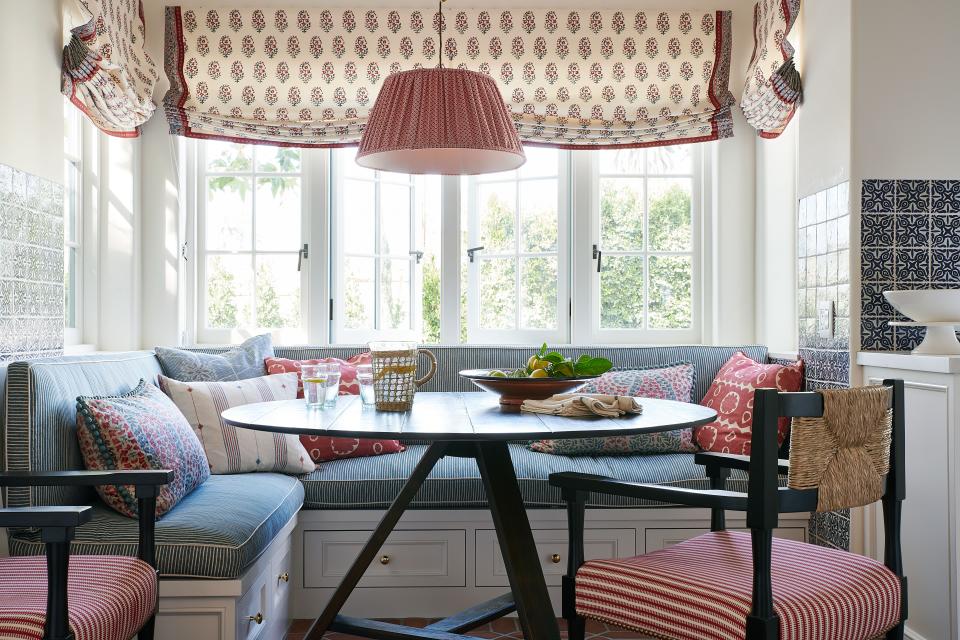 Roman shades by Hill Brown dress the windows in a sunny breakfast nook, where competing fabrics take on a life of their own. A Soane ceiling light coordinates with the rosy ticking stripe, also by Soane, on the Hollywood at Home chairs, while the Fermoie-upholstered banquette is punctuated with pillows covered in fabrics by Lisa Fine Textiles, William Laman, Jasper, and Lee Jofa.