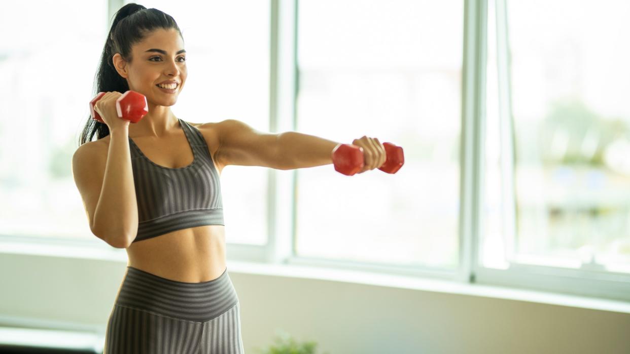  Woman holding a set of dumbbells left arm extended at shoulder height and right arm by head during dumbbell arm workout in studio. 