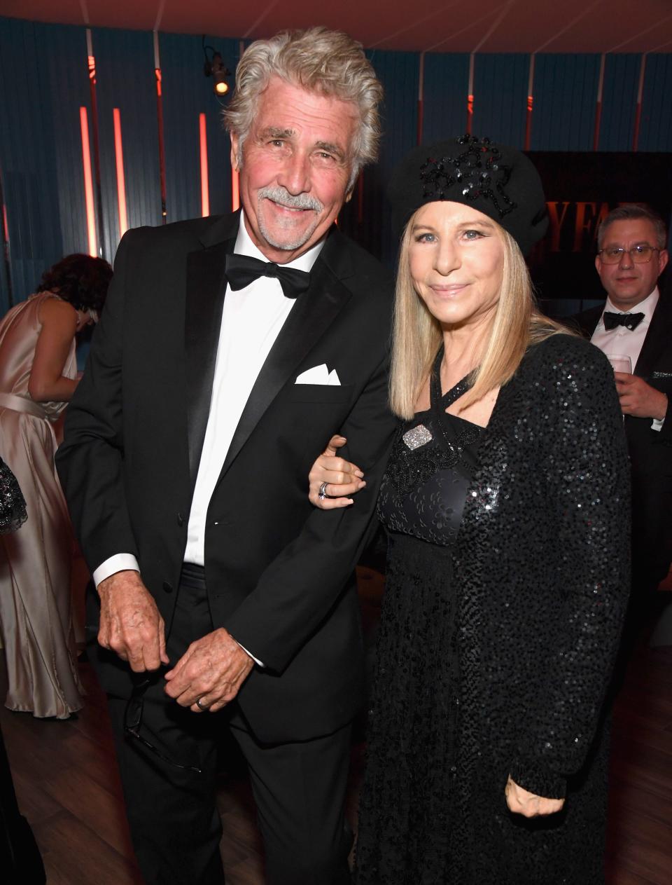 James Brolin, in a tuxedo, poses with Barbra Streisand, in a black beaded dress and matching jacket and hat, at a party.