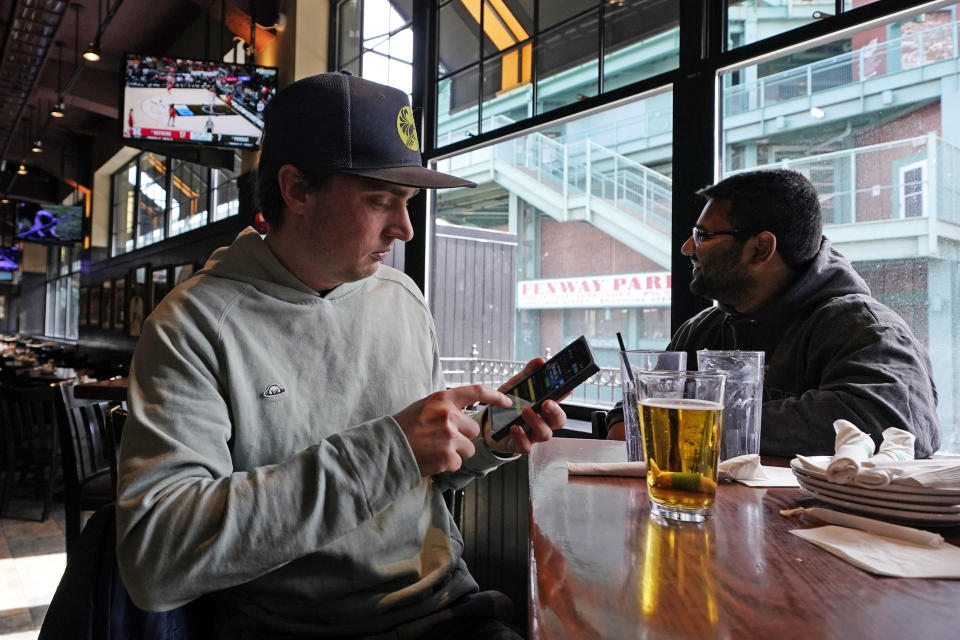Taylor Foehl, left, of Boston, looks at the mobile betting app on his phone after placing a wager, while watching on a men's college basketball game at the Cask 'N Flagon sports bar, Friday, March 10, 2023, near Fenway Park in Boston. Massachusetts sports fans raced to their cell phones Friday, March 10 to begin placing bets as the state allowed online sports wagering just days ahead of tip-off of the NCAA Tournament next week. (AP Photo/Charles Krupa)