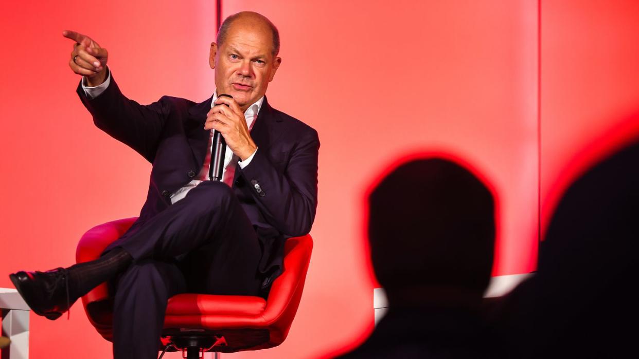 Olaf Scholz hält sitzend ein Mikrofon in der Hand und zeigt mit der rechten Hand auf. 