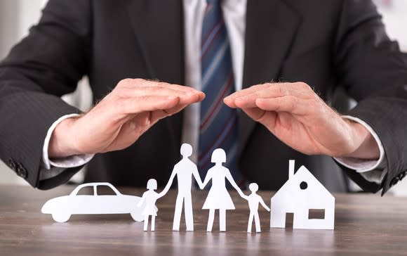 A man in a suit holding his hands over paper cutouts of a family, a car, and a house.