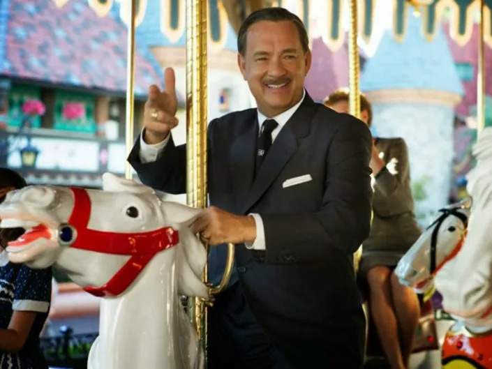 Tom Hanks dressed in a suit and with a mustache giving the two finger point while riding on a carousel.