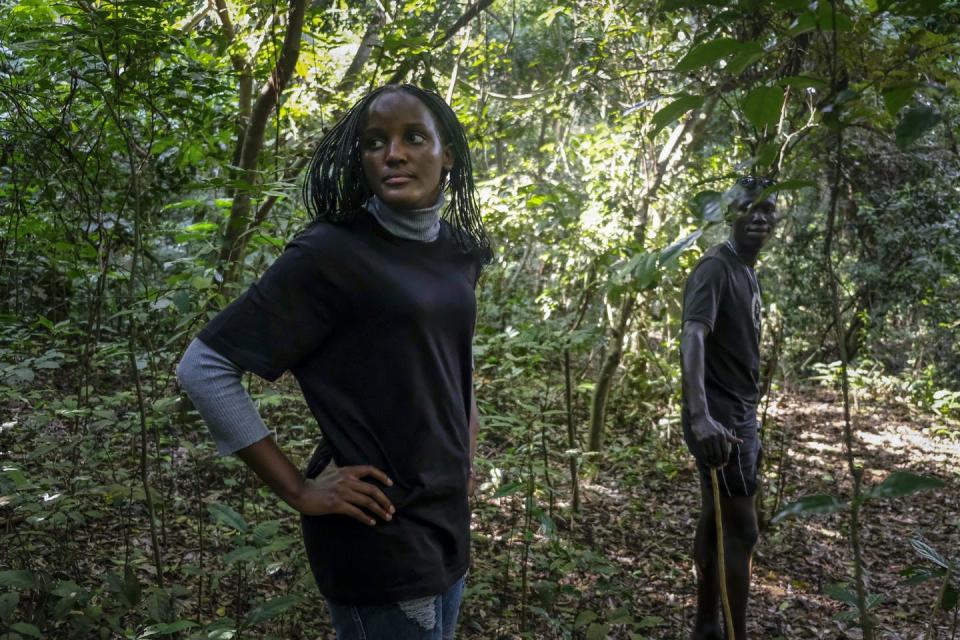 A woman stands in a forested area looking back, past the camera, over her shoulder. A man stands several metres in front of her, also looking back.