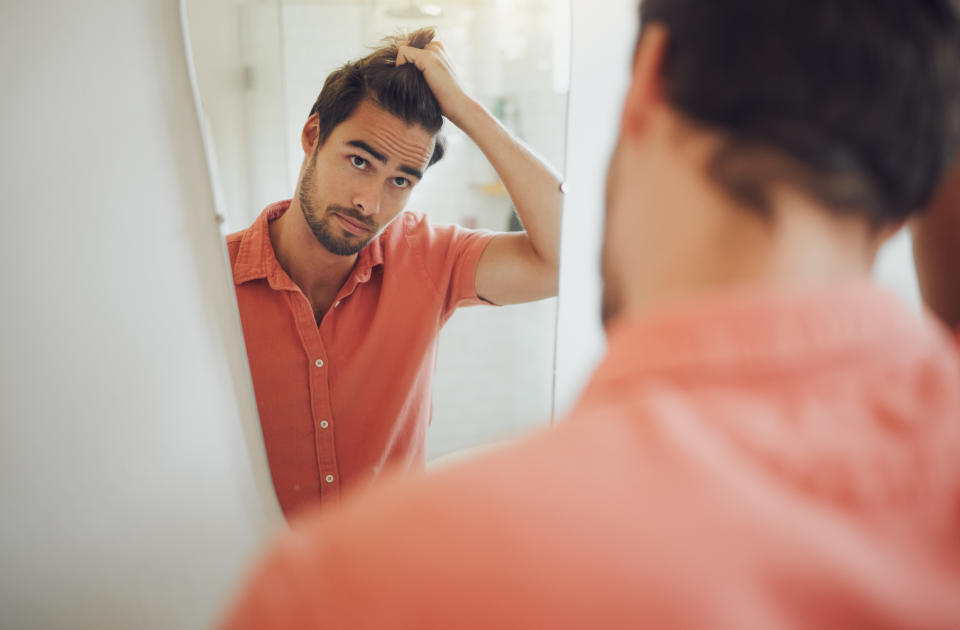 Handsome young caucasian man touching his hair and looking in the bathroom mirror. Male pulling his hair and thinking of getting a haircut. Concerned man worried about dandruff, receding hairline or hair loss