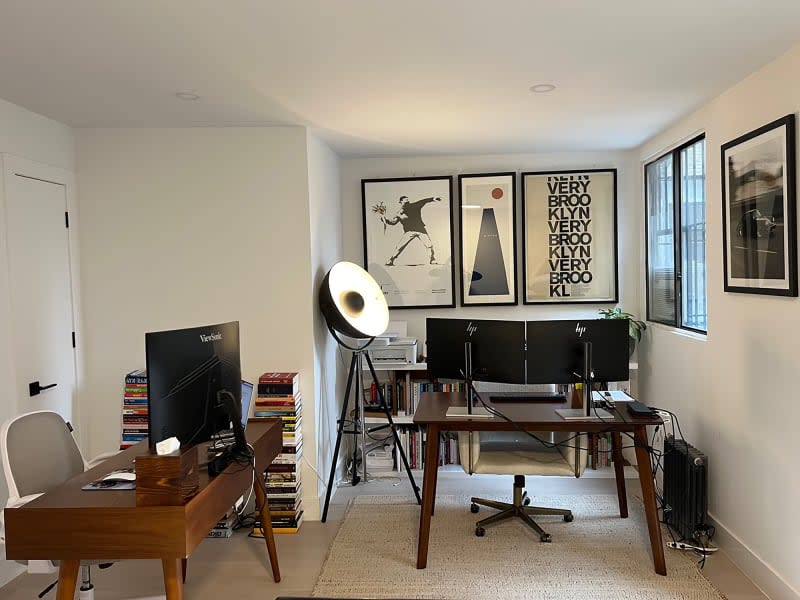 Two wooden desks, stacked books, and bookcases in neutral office with artwork hung on walls.