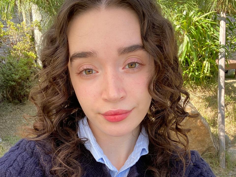 Selfie of the writer in a blue collared shirt, dark-blue cardigan, and minimal makeup. The writer smiles softly and stands in front of green trees