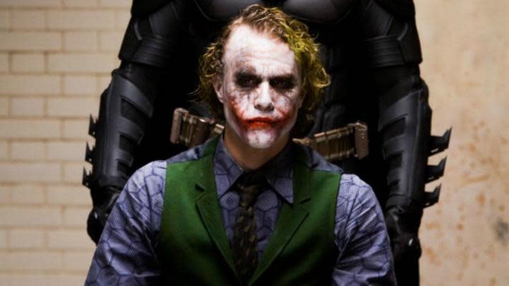 Heath Ledger as the Joker sitting down with Batman behind him in The Dark Knight.